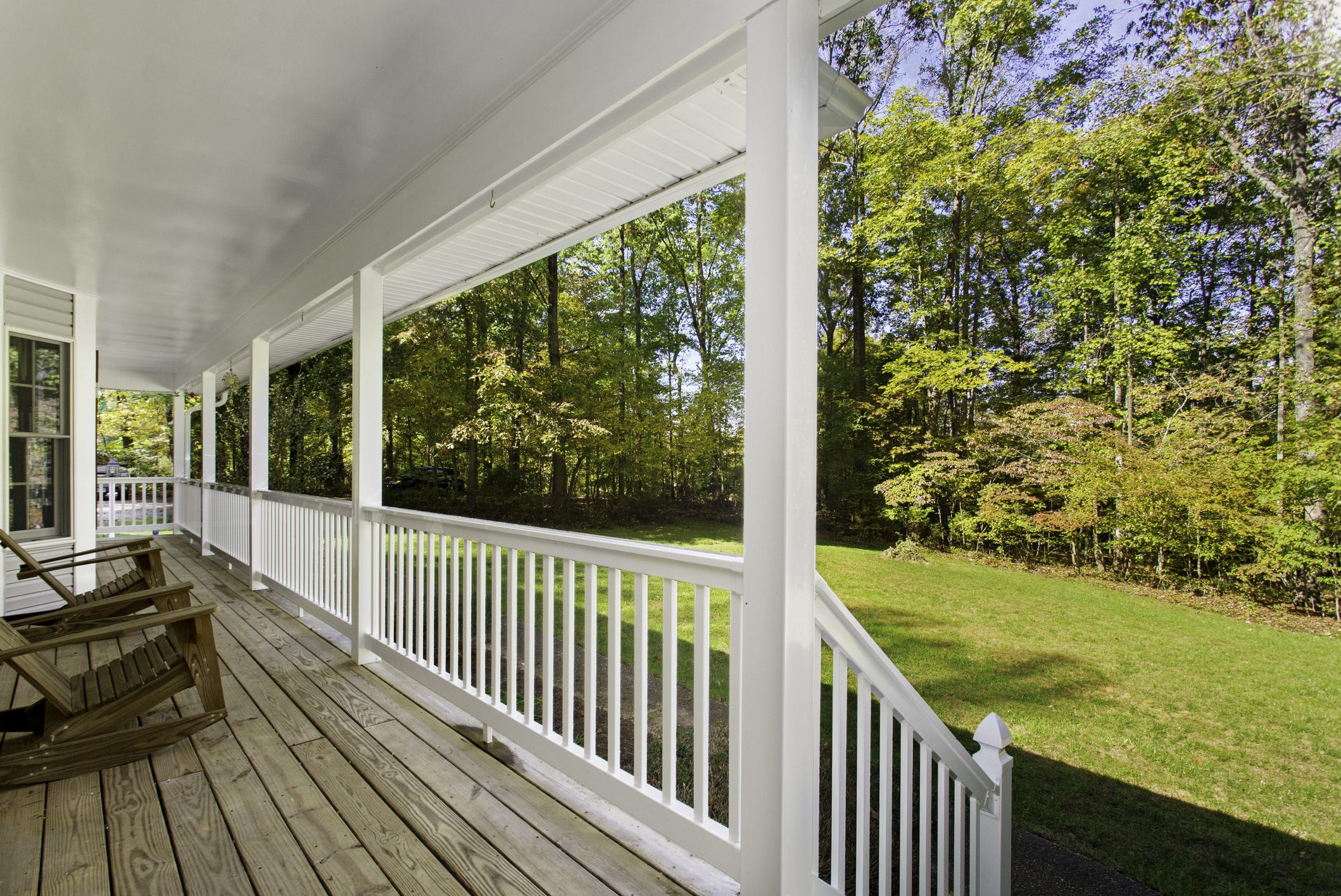 This enchanting front porch features wooden flooring and white railings, offering a picturesque view of lush green grass and tall trees. The inviting, open space is perfect for relaxation and unwinding, creating a peaceful atmosphere for homeowners and visitors.