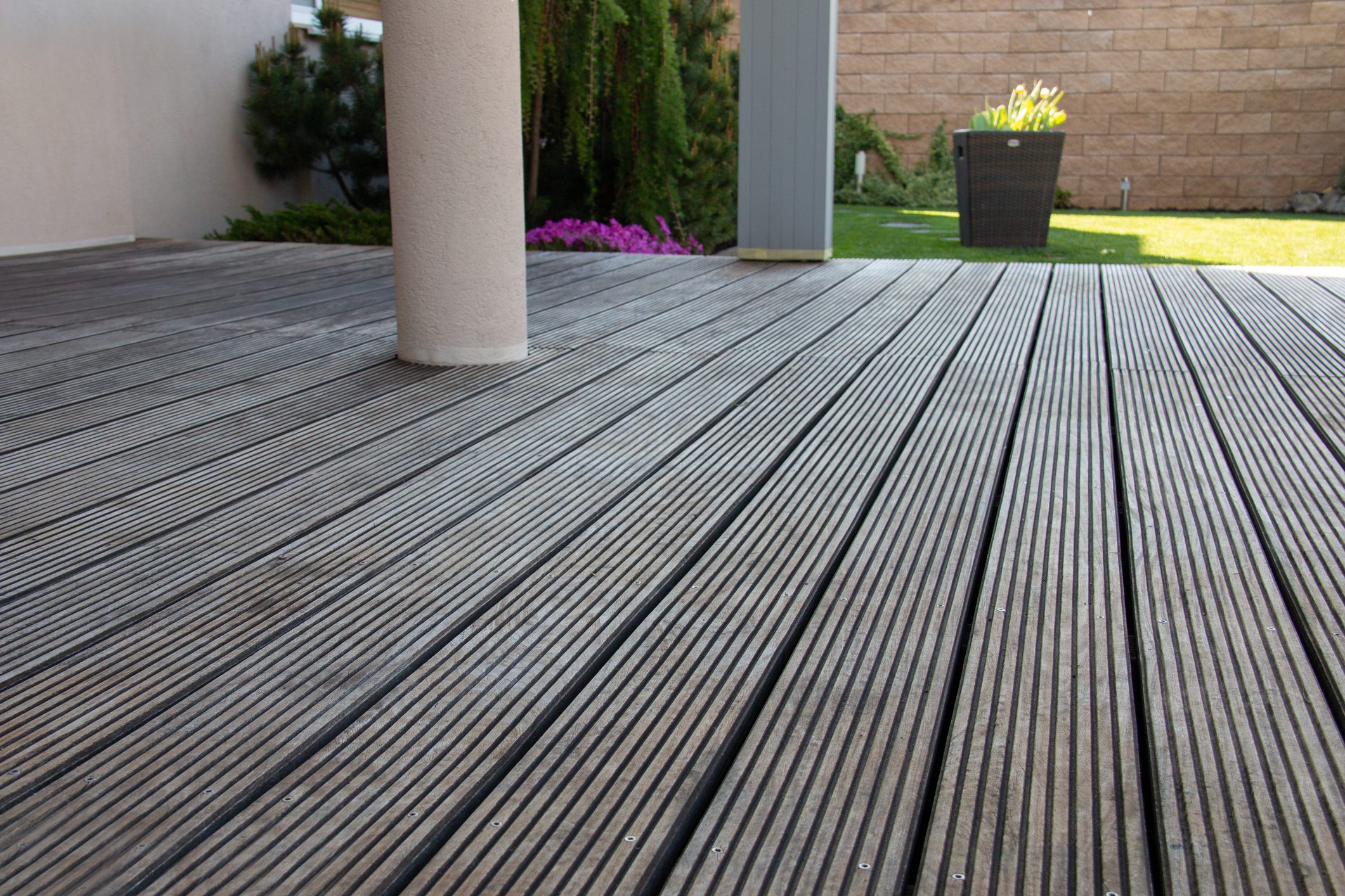 A well-maintained wooden deck showcases horizontal planks in a sunny outdoor area. Lush plants and a small flower bed enhance the serene atmosphere of the space.